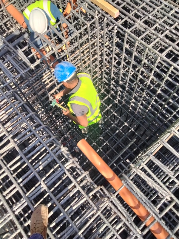 Steel fixer in a blue hard hat tying reinforcement inside a deep rebar cage, with a second worker above